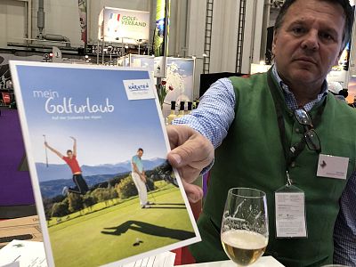 Man holding a golf brochure at a trade show, featuring a joyful golfer on the cover and a glass of beer in the foreground.