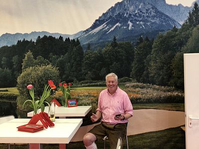 A man in a pink shirt sits at a cafe table with red flowers. Behind him is a mural of a mountain and forest scene.