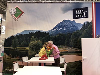 A couple kisses in front of a backdrop featuring mountains and a golf course, with a sign reading 'Golf Tirol Card'. They are at a travel expo.