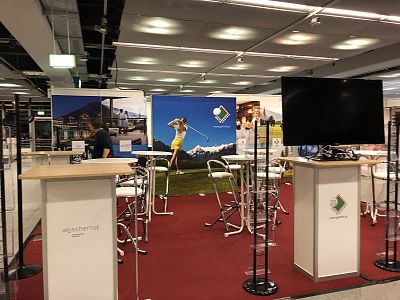 Exhibition booth with high tables, chairs, and a golf-themed backdrop in a spacious hall with a red carpet flooring.