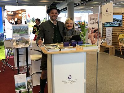 Man in traditional attire and woman smiling at a promotional booth. Flowers and brochures are on the counter at a trade fair.
