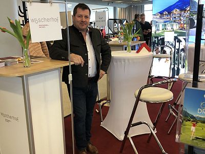 Man in business attire stands at a trade booth with modern decor, tulip flowers, and promotional materials, indicating a professional exhibition setting.