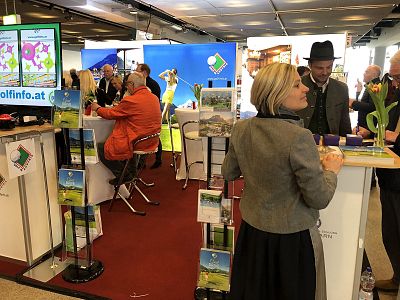 People interacting at a busy exhibition booth with colorful displays, brochures, and digital screens promoting travel destinations.