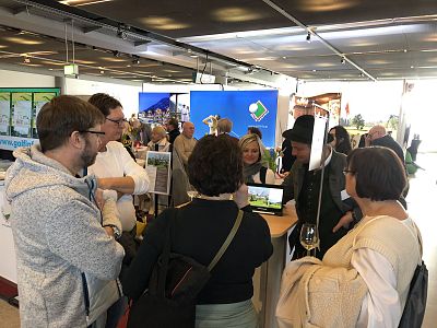 A group of people interact at a trade fair, engaging with a display. The setting is bustling, reflecting a lively and interactive environment.