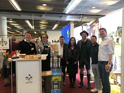 Eine Gruppe von sieben Personen steht lächelnd in einer Halle. Neben einem Messestand mit kulinarischen Produkten tragen einige traditionelle bayerische Kleidung. Ein Banner ist sichtbar.