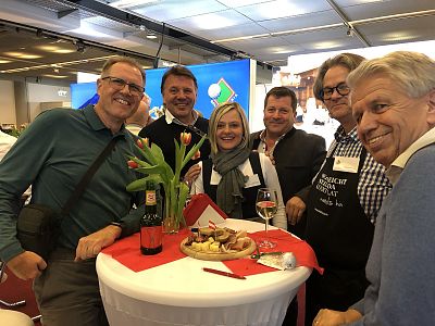 A group of six people smiling and standing around a small table with drinks and food at an indoor event. Tulips decorate the table, creating a cheerful ambiance.