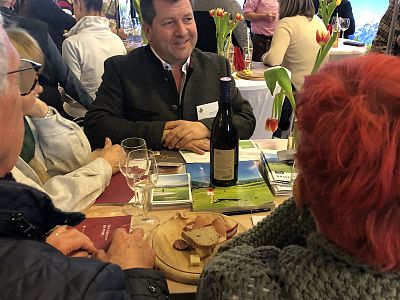 Group of people sitting at a table with wine bottles, glasses, and tulips, engaged in conversation. A cozy, informal setting with snacks and brochures.