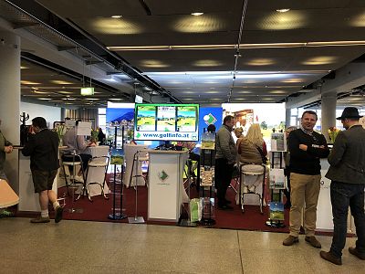 Exhibition booth with interactive displays and visitors engaging, located in a contemporary event space with overhead lighting.