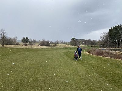 Person pushing a golf cart on a green course under cloudy skies, surrounded by sparse trees and light snowfall, suggesting a cold climate.