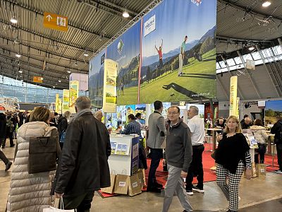 People walking through a trade show with booths displaying travel information. Bright banners and displays create a lively atmosphere in the spacious hall.
