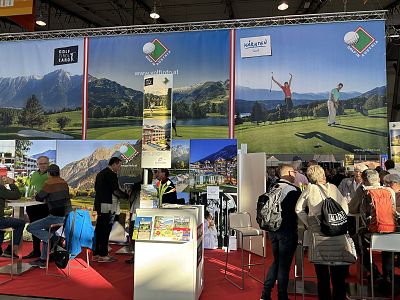 A busy expo booth with large banners of mountains and golf courses, showcasing tourist destinations, while people browse brochures and information on tables.