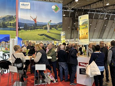 People gather at a golf expo, engaging with displays and each other. The venue has high ceilings and banners, creating an energetic atmosphere.