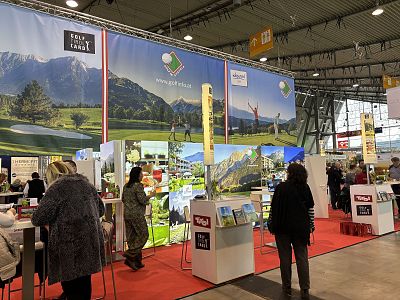 People at a travel trade show, exploring booths with posters and brochures under bright lighting in a spacious exhibition area.
