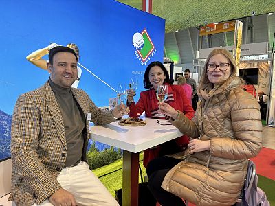 Drei Personen sitzen an einem Tisch auf einer Messe, lächeln und stoßen mit Weingläsern an. Im Hintergrund sieht man einen großen Bildschirm mit Golfmotiv.