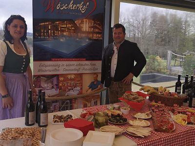 Two people in traditional attire stand by a display of food and wine at an event. A banner features Wöscherhof, with plates of appetizers on a red-checked table.