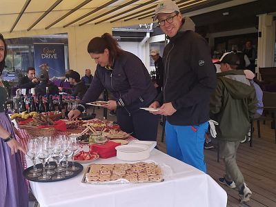 People at an outdoor event enjoying a buffet with assorted food, wine bottles, and cookies under a shaded terrace.