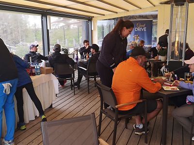 People sitting and standing in a casual dining area, some wearing golf attire, enjoying food and drinks together indoors.