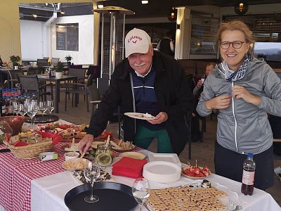 Two people at a buffet table outdoors, covered with a checkered cloth, offering various foods and drinks. They're smiling and appear to enjoy the setting.