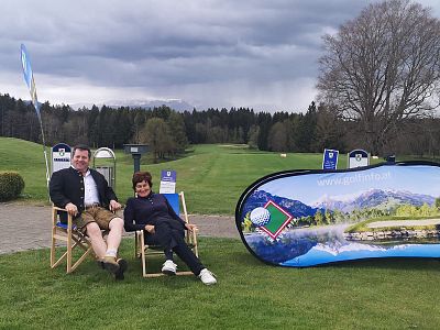 A man and woman sit on chairs on a golf course, smiling. A promotional banner with a mountain scene is nearby, under a cloudy sky.
