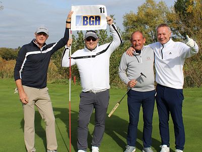 Four men are posing on a golf course, smiling at the camera. One is holding a "BGA" sign. They are dressed in golf attire and standing on a green.