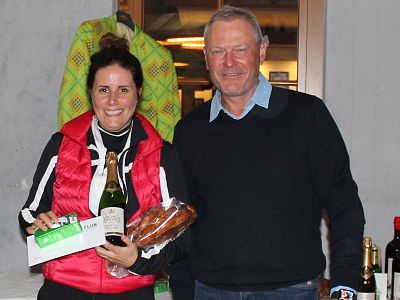 A woman and a man standing together, smiling, holding a bottle of wine and a gift package. They appear to be celebrating in a casual indoor setting.