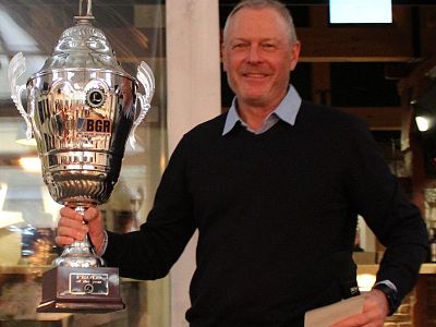 Ein Mann in einem dunkelblauen Pullover hält einen großen Siegerpokal in der Hand und lächelt. Der Hintergrund zeigt ein hell erleuchtetes Gebäudeinnere.
