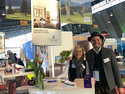 Two people stand smiling at a booth with a banner showing a hotel and golf course. The booth has tulips and promotional materials. The setting is a trade show.