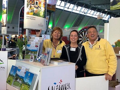 A group of three people smiling at a trade show booth, surrounded by brochures and a dartboard, wearing branded yellow jackets.