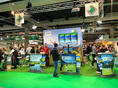 A trade fair booth with people interacting around promotional displays, featuring colorful banners and digital kiosks, emphasizing travel and tourism.