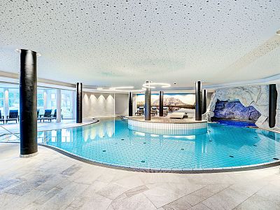 Indoor swimming pool with black columns, a scenic wall mural, and a cave-like area with a waterfall, surrounded by a light-filled and luxurious interior.