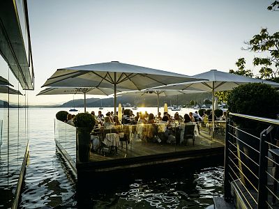 People dining at sunset on a lakeside patio with large umbrellas, reflecting water, and soft evening light.