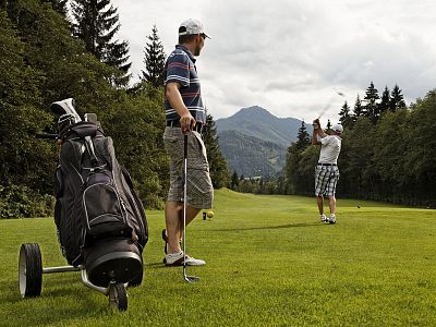 Two golfers on a lush green course with mountains and trees in the background. One prepares to swing, the other observes. A golf bag rests nearby, under a cloudy sky.
