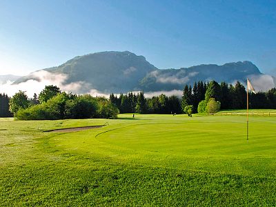 Grüner Golfplatz unter klarem Himmel, umgeben von Bäumen und Bergen. Nebelschwaden ziehen über die Landschaft, die von Sonnenlicht erhellt wird.