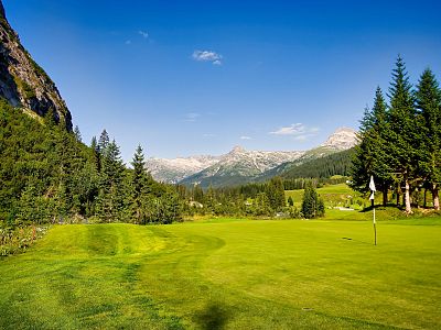 A scenic golf course with lush greens, surrounded by pine trees and mountains under a clear blue sky, offering a tranquil and picturesque landscape.