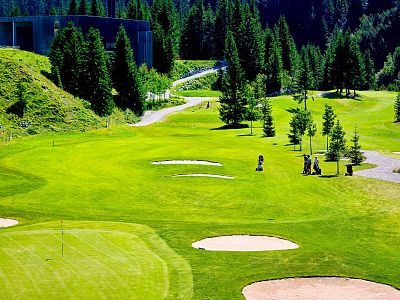 A vibrant golf course with green hills, tall trees, and golfers under bright sunlight. The scene is peaceful and serene.