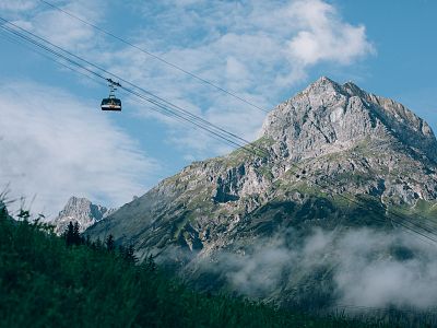 Eine Seilbahn schwebt vor einem imposanten Bergmassiv unter bewölktem Himmel. Im Vordergrund sieht man grüne Hänge und vereinzelte Wolkennebel.