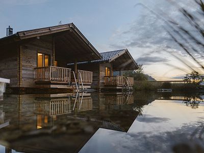 Wooden cabins by a lake at sunset, reflecting on water, with gentle lighting and a peaceful sky, creating a tranquil and serene atmosphere.