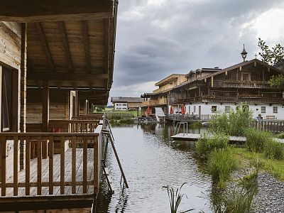 Wooden houses with balconies by a peaceful lake, surrounded by greenery under a cloudy sky.