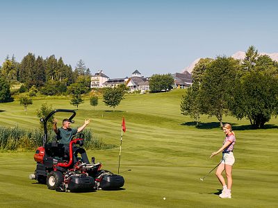 A person on a lawnmower gestures to a golfer holding a putter on a green golf course, with a clubhouse and trees in the background.