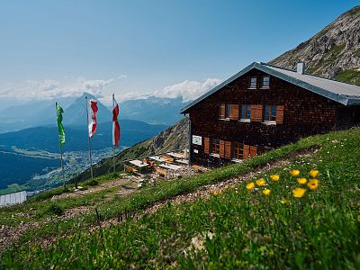 Mountain hut on a hillside with flags, overlooking a valley and distant mountains under a clear blue sky. Green grass and yellow flowers in the foreground.