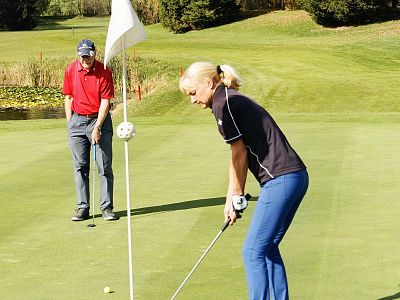 A woman putting on a golf green while a man watches, under clear skies. The scene is vibrant with lush greenery around, capturing a sunny day outdoors.