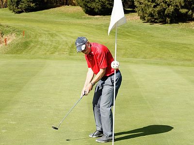 A golfer in a red shirt putts on a sunny green, with a white flagstick nearby. The lush grass and clear sky create a peaceful golfing atmosphere.