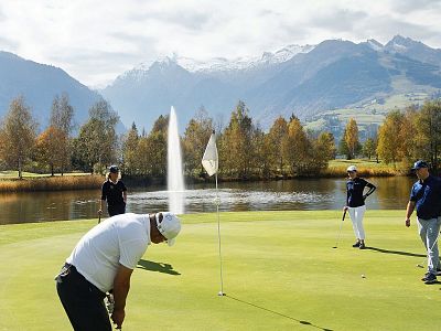 People playing golf on a green near a lake with mountains in the background. A fountain sprays water, and autumn trees line the course under a blue sky.