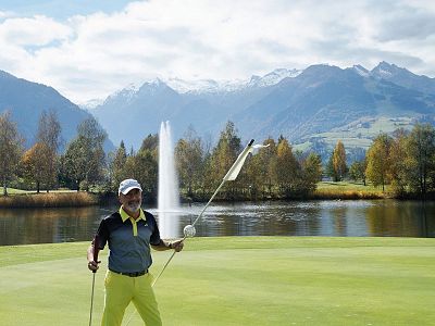 A golfer on a green near a lake with a fountain, snowy mountains and a cloudy sky in the background.