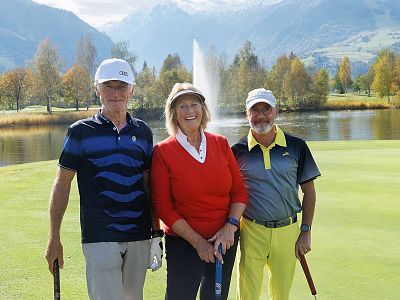 Drei Personen stehen auf einem Golfplatz vor einem Teich mit einem Springbrunnen. Im Hintergrund sind Bäume mit Herbstlaub und Berge zu sehen.