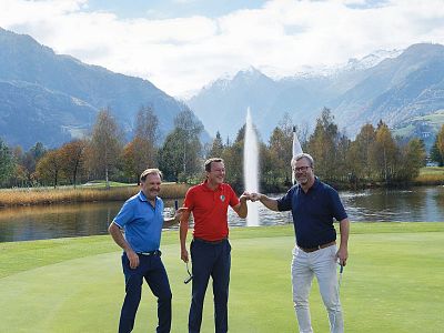 Three people on a golf course surrounded by mountains and a lake, enjoying a sunny day with autumn trees and water fountains in the background.