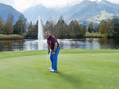 A man stands on a golf green, putting with a club. Behind him, a lake, trees, a fountain, and mountains under a clear sky create a picturesque scene.