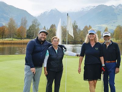 Vier Personen stehen auf einem Golfplatz vor einer Bergkulisse. Zwei von ihnen halten Golfschläger, und ein Fahnenmast ist in der Mitte sichtbar.