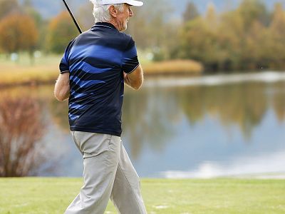 A man in a blue and black shirt and gray pants playing golf at a scenic course. He's mid-swing near a lake with trees displaying autumn colors.