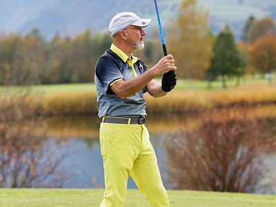Older man playing golf, wearing a cap and yellow pants, swings his club on a lush course with autumn trees and a pond in the background.